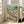 May include: Framed poster print in light wood frame leaning against white wall next to beige armchair. Artwork shows two cartoon ducks doing yoga on beach - one in colorful floral leggings in tree pose, another in striped shirt holding wine glass. Beach scene with palm tree, ocean waves, yoga mat, wine bottles and snacks scattered on sand. Vintage-style illustration with "DUCK YOGA" text at top in multicolored letters.