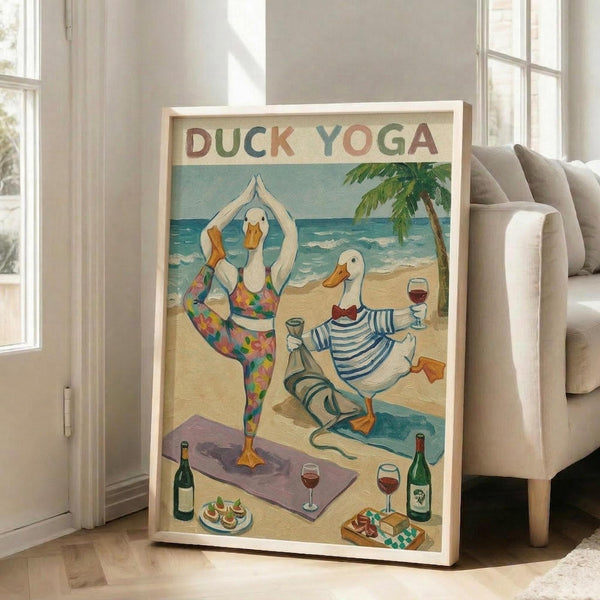 May include: Framed poster print in light wood frame leaning against white wall next to beige armchair. Artwork shows two cartoon ducks doing yoga on beach - one in colorful floral leggings in tree pose, another in striped shirt holding wine glass. Beach scene with palm tree, ocean waves, yoga mat, wine bottles and snacks scattered on sand. Vintage-style illustration with "DUCK YOGA" text at top in multicolored letters.