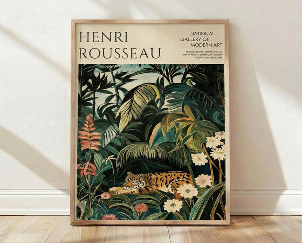 May include: Framed poster print featuring Henri Rousseau's jungle artwork with a resting tiger among lush tropical foliage in deep greens, yellows, and coral tones. White flowers and palm fronds surround the spotted big cat. The cream-colored poster displays "HENRI ROUSSEAU" and "NATIONAL GALLERY OF MODERN ART" text at top. Light wood frame sits against white wall on wooden floor with natural lighting casting soft shadows.