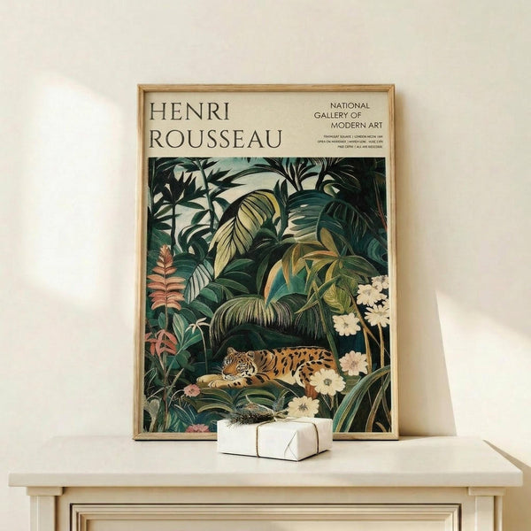 May include: Framed poster featuring Henri Rousseau's jungle scene with a resting tiger among lush tropical foliage in deep greens, displayed on a light wooden shelf against a cream wall. The artwork shows dense palm fronds, exotic plants, and white flowers surrounding the spotted big cat. A small white gift box sits beside the natural wood frame.