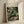 May include: Framed art print leaning against white textured wall with natural light casting diagonal shadows. The poster features a lush jungle scene with dense green tropical foliage, palm fronds, and colorful flowers surrounding a resting leopard with spotted golden coat. Text reads "Henri Rousseau" and "National Gallery of Modern Art" at top. Light wooden frame against neutral background creates warm, gallery-style display.