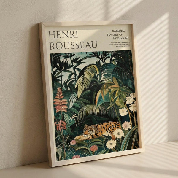 May include: Framed art print leaning against white textured wall with natural light casting diagonal shadows. The poster features a lush jungle scene with dense green tropical foliage, palm fronds, and colorful flowers surrounding a resting leopard with spotted golden coat. Text reads "Henri Rousseau" and "National Gallery of Modern Art" at top. Light wooden frame against neutral background creates warm, gallery-style display.