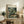 May include: Framed Henri Rousseau jungle art print displayed on rustic wooden sideboard in warm-toned living room. The botanical poster features a leopard resting among lush tropical foliage with white flowers. Scene shows cream-colored armchair, fern plant, ceramic vases, books, and neutral wall creating cozy home decor setting.