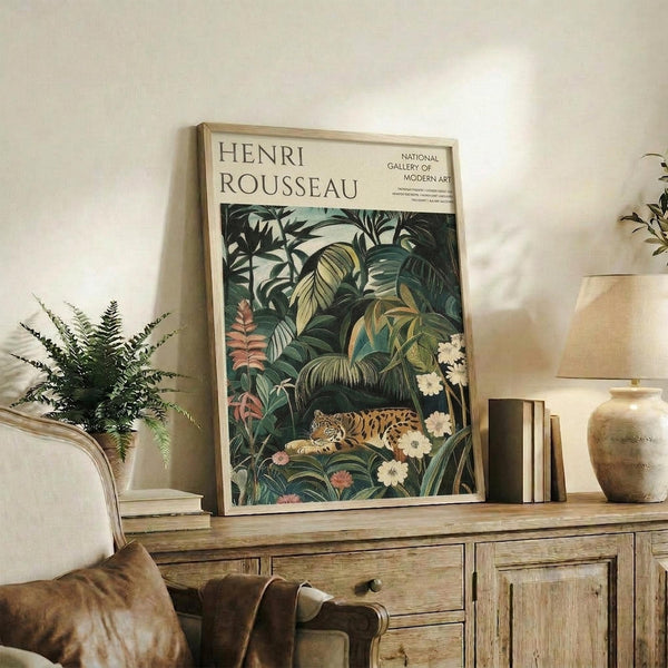 May include: Framed Henri Rousseau jungle art print displayed on rustic wooden sideboard in warm-toned living room. The botanical poster features a leopard resting among lush tropical foliage with white flowers. Scene shows cream-colored armchair, fern plant, ceramic vases, books, and neutral wall creating cozy home decor setting.