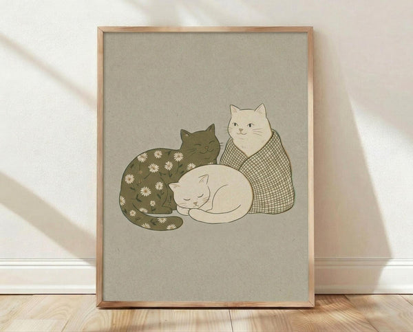 May include: Framed poster print leaning against white wall on light wooden floor with natural sunlight casting shadows. The artwork shows three cats in muted earth tones - a dark green cat with white daisy pattern sleeping, a cream cat with crosshatch pattern sitting upright, and a small white kitten curled up between them. The illustration has a soft, hand-drawn style on beige textured background within a natural wood frame.