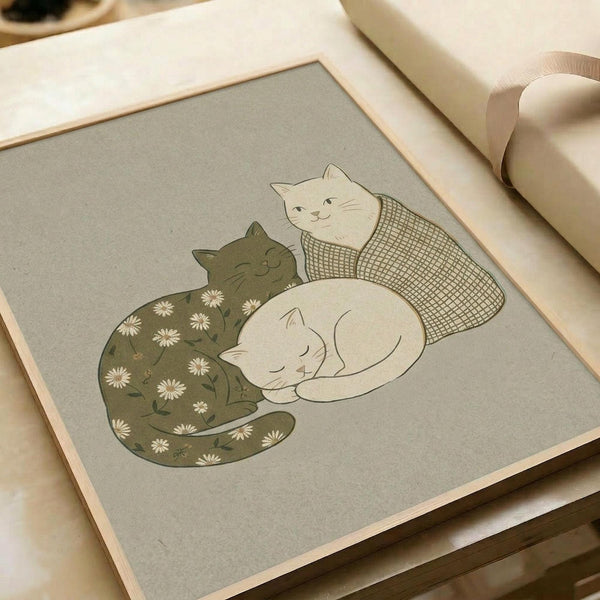 May include: Framed art print on gray paper showing three sleeping cats cuddled together. One olive green cat with white daisy pattern, one cream-colored cat with checkered pattern, and one solid white cat. Print has wooden frame and sits on white surface with rolled paper in background. Soft, minimalist illustration style with muted earth tones.