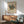 May include: Framed Kandinsky abstract art poster hanging above wooden credenza in modern living room. Colorful geometric composition with circles, triangles, and lines on cream background. Mid-century furniture includes sage green upholstered chair with wooden arms, round coffee table, and white ceramic lamp. Natural sunlight creates shadows on white wall.