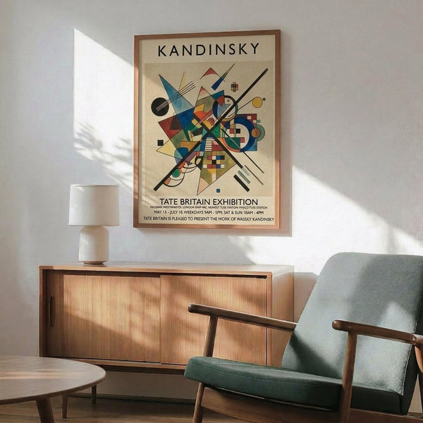 May include: Framed Kandinsky abstract art poster hanging above wooden credenza in modern living room. Colorful geometric composition with circles, triangles, and lines on cream background. Mid-century furniture includes sage green upholstered chair with wooden arms, round coffee table, and white ceramic lamp. Natural sunlight creates shadows on white wall.