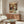 May include: Vintage-style poster showing two white ducks with orange beaks in formal attire sharing a romantic dinner in an ornate room with chandelier, displayed above a walnut credenza with brass hardware in a modern living room with cream walls, white curved chair, and oval glass coffee table