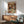 May include: Vintage-style poster featuring two white ducks with orange beaks dressed formally at an elegant dinner table, surrounded by wine, chocolates, and roses in an ornate room with chandelier. Shown framed in thin wood frame above mid-century modern credenza next to mint green upholstered armchair and white ceramic lamp in bright, naturally-lit living room.