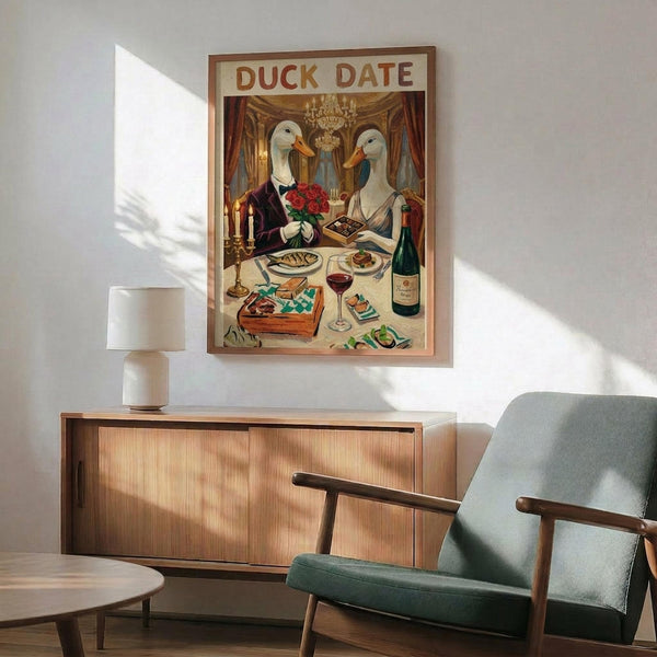 May include: Vintage-style poster featuring two white ducks with orange beaks dressed formally at an elegant dinner table, surrounded by wine, chocolates, and roses in an ornate room with chandelier. Shown framed in thin wood frame above mid-century modern credenza next to mint green upholstered armchair and white ceramic lamp in bright, naturally-lit living room.