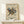 May include: Framed Kandinsky abstract art poster featuring geometric shapes, lines, circles and triangles in blue, red, yellow and green colors on cream background. Exhibition poster text reads "Tate Britain Exhibition" with venue details. Displayed on light wood shelf against white wall next to small wrapped gift box. Natural lighting creates soft shadows on minimalist home decor setting.