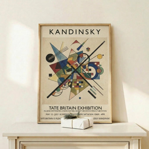 May include: Framed Kandinsky abstract art poster featuring geometric shapes, lines, circles and triangles in blue, red, yellow and green colors on cream background. Exhibition poster text reads "Tate Britain Exhibition" with venue details. Displayed on light wood shelf against white wall next to small wrapped gift box. Natural lighting creates soft shadows on minimalist home decor setting.