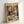 May include: Framed vintage-style poster print leaning against white wall corner. Features two white ducks with orange bills dressed formally - one in burgundy suit with bow tie holding red roses, other in white dress. They sit at elegant dining table with wine bottle, glasses, candles, chocolates, and various dishes. Ornate restaurant interior with crystal chandeliers and draped curtains in background. Warm golden and brown color palette in decorative illustration style.