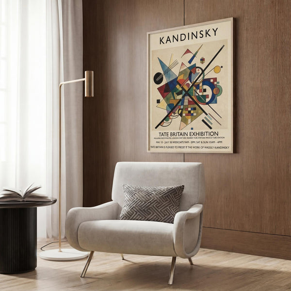 May include: Modern living room with cream armchair and geometric pillow, brass floor lamp, dark wood paneling, and framed abstract art poster featuring colorful geometric shapes, circles, and lines in blues, reds, and yellows with "KANDINSKY" and "TATE BRITAIN EXHIBITION" text on cream background.
