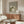 May include: Framed poster showing three stylized cats in muted green, white and beige tones - one cat with daisy pattern, one with checkered design, and one solid white cat sleeping. Displayed above a walnut wood credenza in a minimalist living room with cream walls, white upholstered chair, glass coffee table, and modern mushroom table lamp.