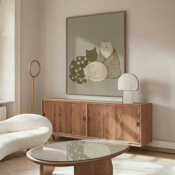 May include: Framed poster showing three stylized cats in muted green, white and beige tones - one cat with daisy pattern, one with checkered design, and one solid white cat sleeping. Displayed above a walnut wood credenza in a minimalist living room with cream walls, white upholstered chair, glass coffee table, and modern mushroom table lamp.