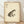 May include: Framed vintage-style illustration of a spotted brown and tan frog with prominent eyes on aged cream paper with Japanese characters and red stamps in upper left corner, displayed in natural wood frame leaning against white wall on light wooden floor