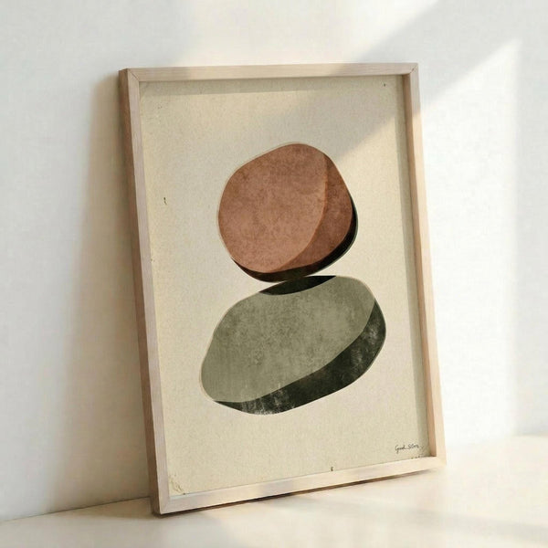 May include: Framed abstract art print featuring two balanced oval stones in earthy terracotta and sage green tones on cream textured paper, displayed in light wooden frame leaning against white wall with natural lighting creating soft shadows