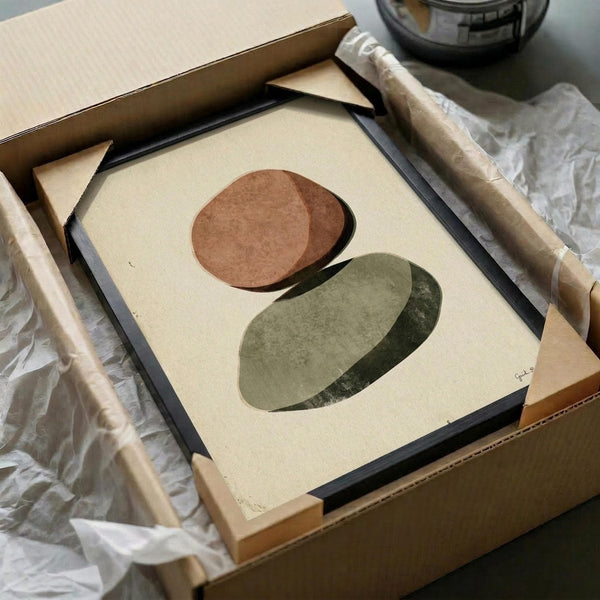 May include: Abstract art print featuring two overlapping oval shapes in earthy tones - a warm terracotta brown oval above and sage green oval below on cream background. Framed artwork shown in black frame being unpacked from cardboard box with protective tissue paper and wooden dowels visible around shipping materials.