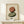 May include: Framed abstract art print featuring two stacked oval stones in terracotta orange and sage green watercolor style, displayed on light wood console table against cream wall. Small wrapped white gift box with evergreen sprig sits beside the natural wood frame. Minimalist Scandinavian home decor styling with soft natural lighting.