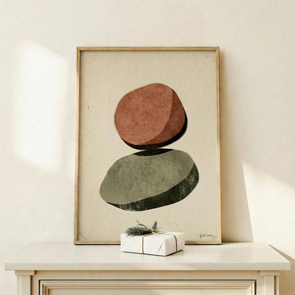 May include: Framed abstract art print featuring two stacked oval stones in terracotta orange and sage green watercolor style, displayed on light wood console table against cream wall. Small wrapped white gift box with evergreen sprig sits beside the natural wood frame. Minimalist Scandinavian home decor styling with soft natural lighting.