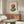 May include: Abstract minimalist poster with stacked oval stones in terracotta and sage green tones, displayed in thin wooden frame above mid-century walnut credenza. Modern living room setting features cream upholstered chair, oval glass coffee table with colorful art book, brass floor lamp, and white table lamp. Neutral walls with natural window light create serene, contemporary interior styling.