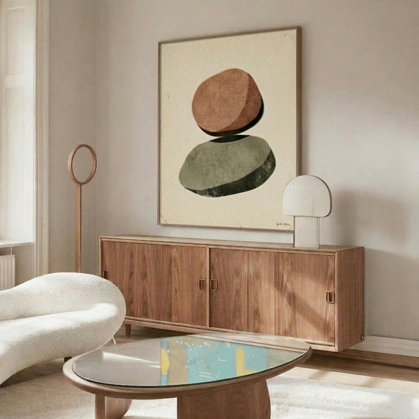 May include: Abstract minimalist poster with stacked oval stones in terracotta and sage green tones, displayed in thin wooden frame above mid-century walnut credenza. Modern living room setting features cream upholstered chair, oval glass coffee table with colorful art book, brass floor lamp, and white table lamp. Neutral walls with natural window light create serene, contemporary interior styling.