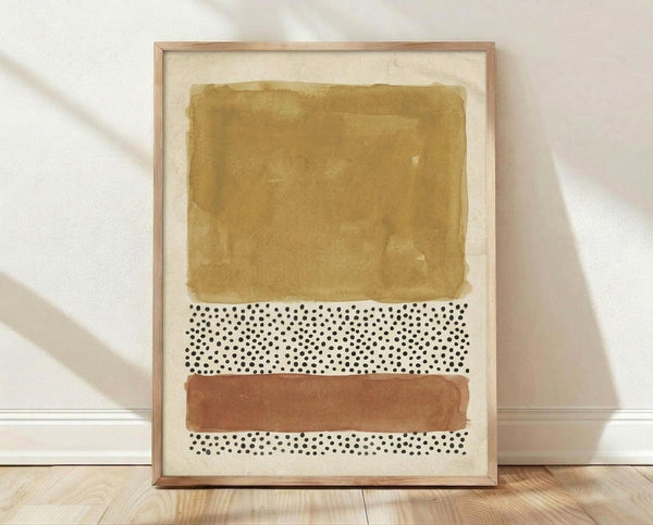 May include: Framed abstract art print leaning against white textured wall on light wood floor. Artwork features earthy ochre watercolor wash at top, black polka dot pattern on cream background in middle, terracotta brown brushstroke band, and smaller dotted section at bottom. Natural wood frame with visible grain. Soft natural lighting creates gentle shadows on wall.