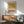 May include: Abstract artwork with earthy ochre brushstroke at top, white section with scattered black dots in middle, and terracotta brushstroke at bottom, displayed in thin wooden frame above mid-century modern wooden credenza. Sage green upholstered armchair with wooden arms sits nearby, white ceramic table lamp on credenza, warm natural light creating shadows on white wall.