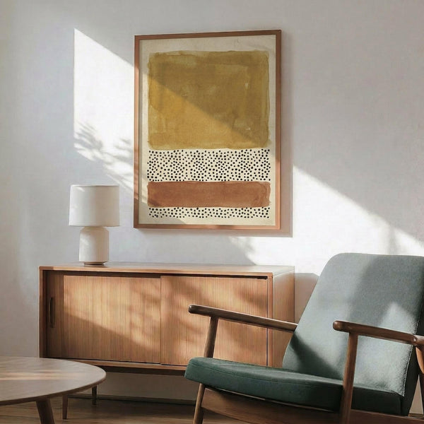 May include: Abstract artwork with earthy ochre brushstroke at top, white section with scattered black dots in middle, and terracotta brushstroke at bottom, displayed in thin wooden frame above mid-century modern wooden credenza. Sage green upholstered armchair with wooden arms sits nearby, white ceramic table lamp on credenza, warm natural light creating shadows on white wall.