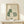 May include: Framed poster print featuring three cats in soft green and cream tones displayed on light wood shelf against white wall. Left cat has green body with white daisy pattern, center cat is small and cream-colored, right cat has green checkered pattern. Small wrapped gift with evergreen sprig sits beside the natural wood frame. Minimalist Scandinavian-style room setting with clean lines and neutral palette.