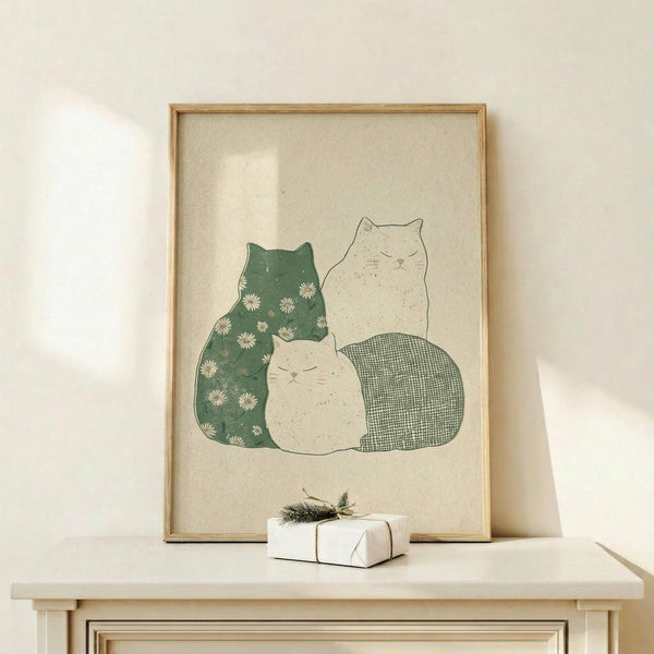 May include: Framed poster print featuring three cats in soft green and cream tones displayed on light wood shelf against white wall. Left cat has green body with white daisy pattern, center cat is small and cream-colored, right cat has green checkered pattern. Small wrapped gift with evergreen sprig sits beside the natural wood frame. Minimalist Scandinavian-style room setting with clean lines and neutral palette.