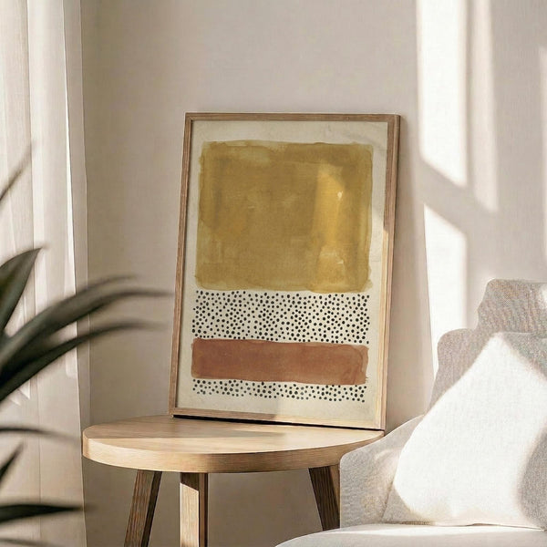 May include: Framed abstract art print with golden ochre watercolor wash at top, white section with black polka dots in middle, and terracotta stripe below, displayed on round wooden side table next to beige upholstered chair with white pillow and plant leaves visible in corner of bright room with natural lighting