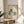May include: Framed art print displayed on round wooden side table next to beige upholstered chair with white pillow. Print shows three stylized cats - one green with white daisy pattern, one white, one smaller white cat, and one with green crosshatch pattern - arranged in sitting positions. Natural wood frame, cream background, soft sunlight streaming across neutral-toned room with plant leaves visible in corner.