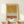 May include: Abstract art print in light wood frame leaning against white wall on wooden shelf. Artwork features layered horizontal sections with golden ochre watercolor wash at top, white section with black polka dots in middle, and terracotta orange watercolor band at bottom with more dotted pattern below. Person's hands visible gripping shelf edge, small wrapped gift with greenery sits nearby. Warm, minimalist styling.