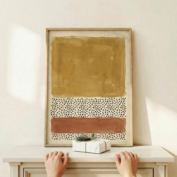 May include: Abstract art print in light wood frame leaning against white wall on wooden shelf. Artwork features layered horizontal sections with golden ochre watercolor wash at top, white section with black polka dots in middle, and terracotta orange watercolor band at bottom with more dotted pattern below. Person's hands visible gripping shelf edge, small wrapped gift with greenery sits nearby. Warm, minimalist styling.