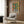 May include: Contemporary living room with beige upholstered armchair and geometric throw pillow. Large framed artwork on wood-paneled wall shows stylized green cactus in black and white striped pot against orange and yellow background. Gold floor lamp and dark round side table with open book complete the modern interior setting.
