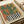 Abstract illustration featuring a stylized green cactus with flowing, curved arms against a warm gradient background of coral, orange and yellow tones. The cactus sits in a black and white striped geometric pot. The artwork has a retro, mid-century modern aesthetic with soft brushstroke textures and earthy colors. Print is displayed on white paper with visible edges, photographed on a light surface next to rolled white paper tubes.