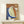 May include: Abstract artwork in wooden frame leaning against white wall on light wood surface, featuring organic curved shapes in terracotta orange, sage green, and navy blue with textured brushstrokes and small cream circle, displayed next to white wrapped gift with evergreen sprig