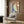 May include: Abstract art print displayed on wood paneled wall above beige upholstered armchair with geometric pillow. Artwork features organic curved shapes in terracotta orange, sage green, and navy blue with textured brushstrokes and small cream circle. Modern brass floor lamp and dark round side table with open book complete the neutral-toned reading nook scene.