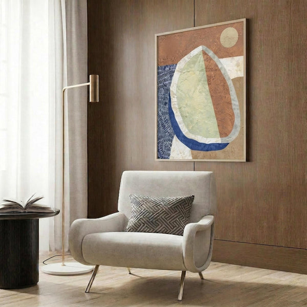 May include: Abstract art print displayed on wood paneled wall above beige upholstered armchair with geometric pillow. Artwork features organic curved shapes in terracotta orange, sage green, and navy blue with textured brushstrokes and small cream circle. Modern brass floor lamp and dark round side table with open book complete the neutral-toned reading nook scene.