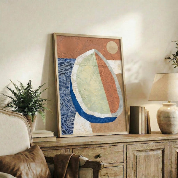 May include: Abstract artwork in light wood frame displayed on rustic wooden sideboard beside fern plant, books, and ceramic vases. Artwork features organic oval shapes in terracotta orange, sage green, and deep blue with textured brushstrokes and small cream circle, set against neutral living room with beige chair and warm lighting.