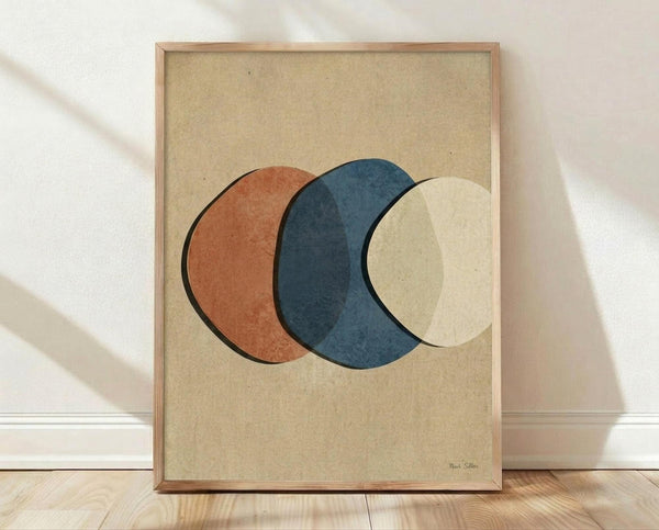 May include: Framed abstract art print featuring three overlapping circles in terracotta orange, deep blue, and cream colors on textured beige background. Displayed in light wood frame leaning against white textured wall above light wooden flooring with natural shadows cast across the scene.