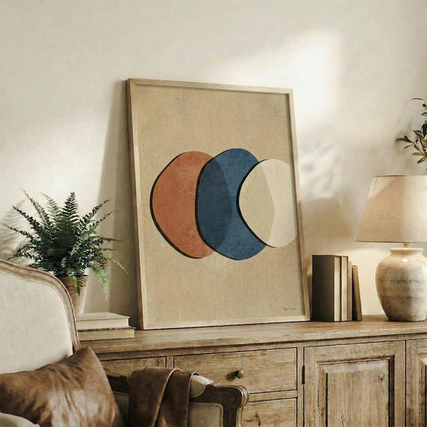 May include: Framed abstract art print featuring three overlapping circles in terracotta orange, navy blue, and cream on beige textured background, displayed on rustic wooden sideboard alongside fern plant, stacked books, and ceramic vases in warm neutral living room setting with white walls and soft natural lighting.