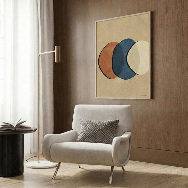 May include: Abstract art print featuring three overlapping circles in orange, navy blue, and cream colors on beige background, displayed above modern beige armchair with geometric patterned pillow. Room setting shows wood paneled wall, brass floor lamp, and dark round side table with open book.