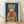May include: Framed art print leaning against white wall on light wood floor. Shows tabby cat wearing mustard yellow hooded raincoat holding red fish, painted in whimsical illustrative style against deep blue background. Natural wood frame with white mat. Soft shadows from window light create gentle lighting across textured white wall.