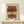 May include: Abstract art print in wooden frame displaying horizontal brushstrokes in terracotta, ochre, and brown tones with central dotted pattern overlay. Artwork sits on light wood console table beside small white wrapped gift with greenery sprig against cream wall.