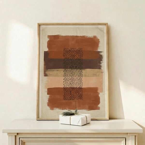 May include: Abstract art print in wooden frame displaying horizontal brushstrokes in terracotta, ochre, and brown tones with central dotted pattern overlay. Artwork sits on light wood console table beside small white wrapped gift with greenery sprig against cream wall.