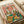 May include: Hands holding a whimsical illustrated poster featuring two white ducks at a picnic. One duck wears sunglasses and a pineapple-print shirt, the other sports a straw hat. The scene includes a wicker basket of yellow ducklings, French bread, wine bottles labeled "Quack Attack" and "Pond Prosecco," cheese board with grapes, and roasted chicken on a green checkered blanket. Pink confetti background with "Duck Picnic" text in colorful letters at top.